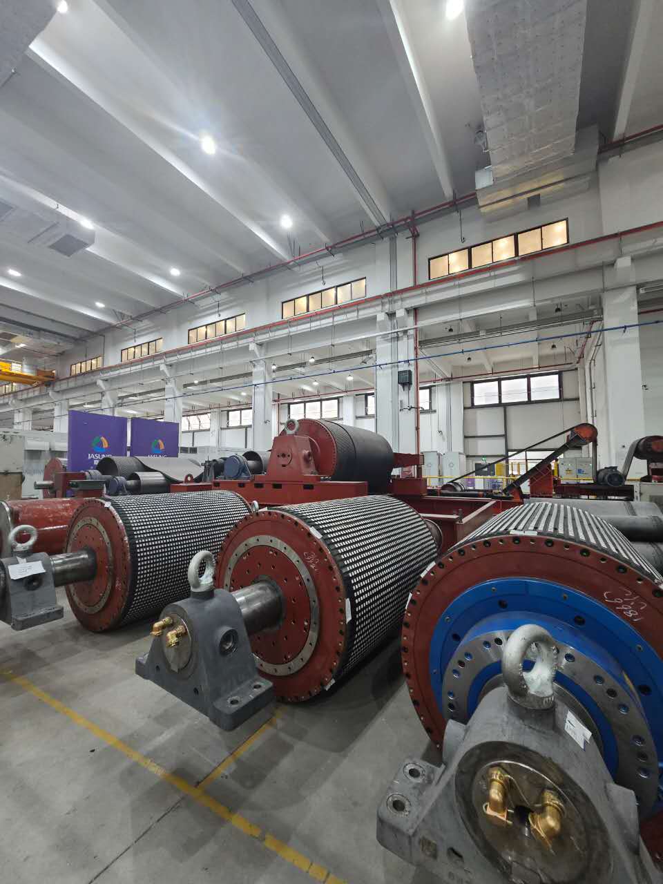 Massive industrial rollers with grid-patterned surfaces lined up inside a bright, spacious manufacturing plant.