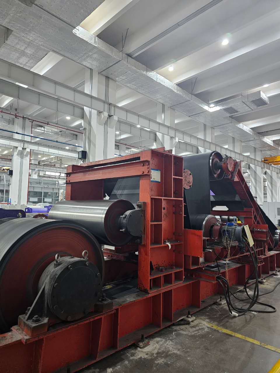 Large industrial machine with red frame and black conveyor belt moving over heavy rollers.