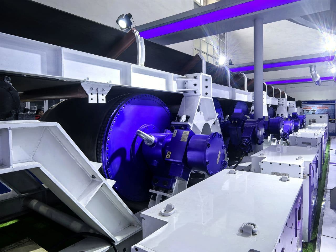 Industrial conveyor belt system with blue motors and white metal frames under purple lighting.
