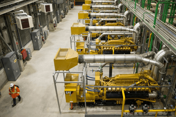 Row of yellow Caterpillar generators in an industrial facility with a worker walking nearby.