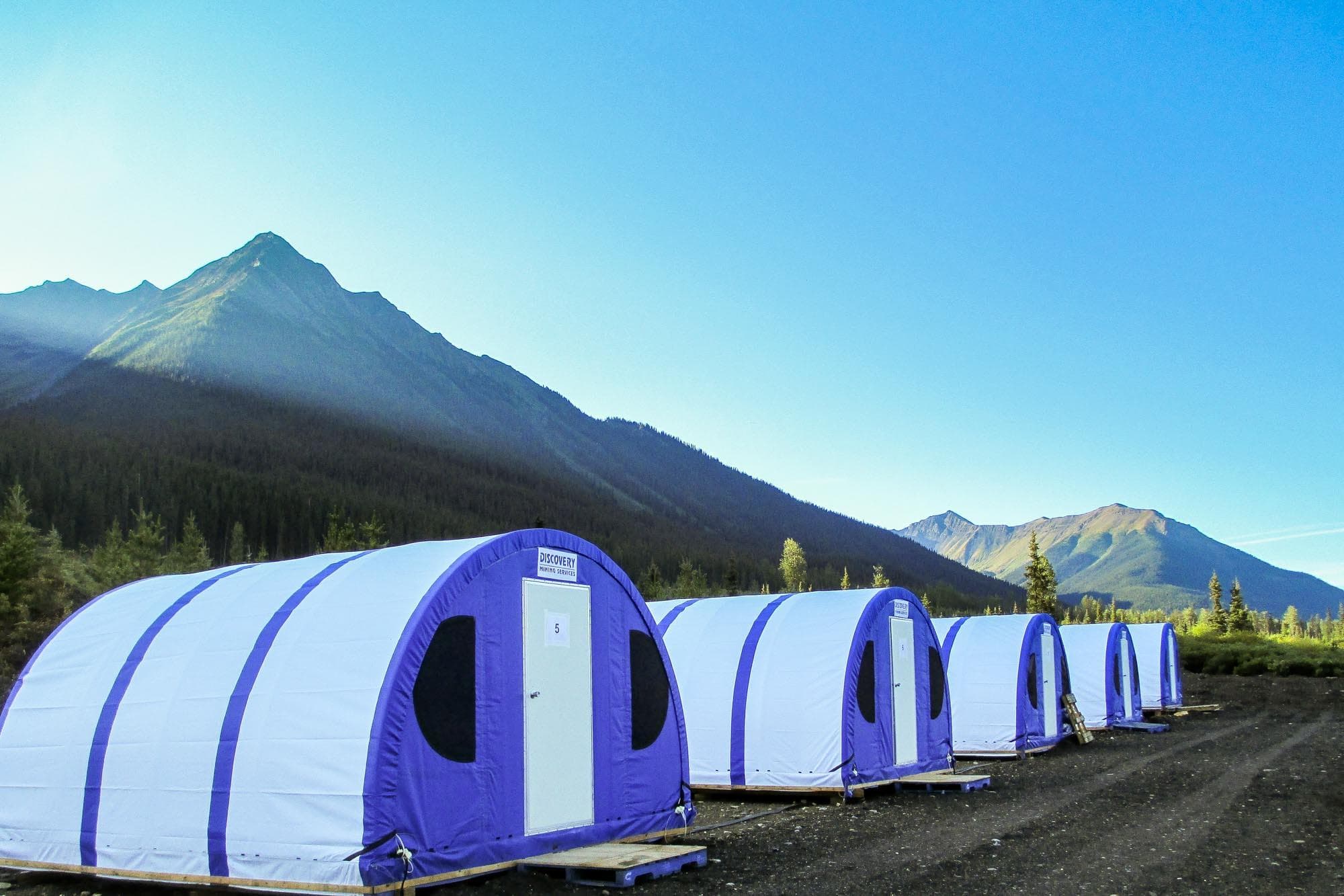White and purple arched tents lined up in a valley below towering, sunlit mountains.