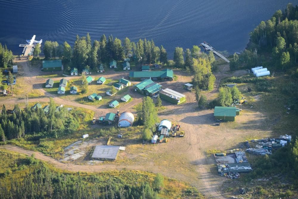 Aerial view of a remote lakeside camp with green-roofed cabins, a floatplane, and a helipad.